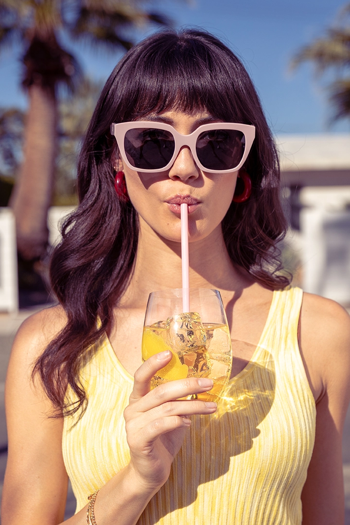 Nikaido Social 3 ‣ Merit/Andrew Woman with purple hair sipping a lemony drink through a pink straw, wearing oversized white sunglasses outdoors.