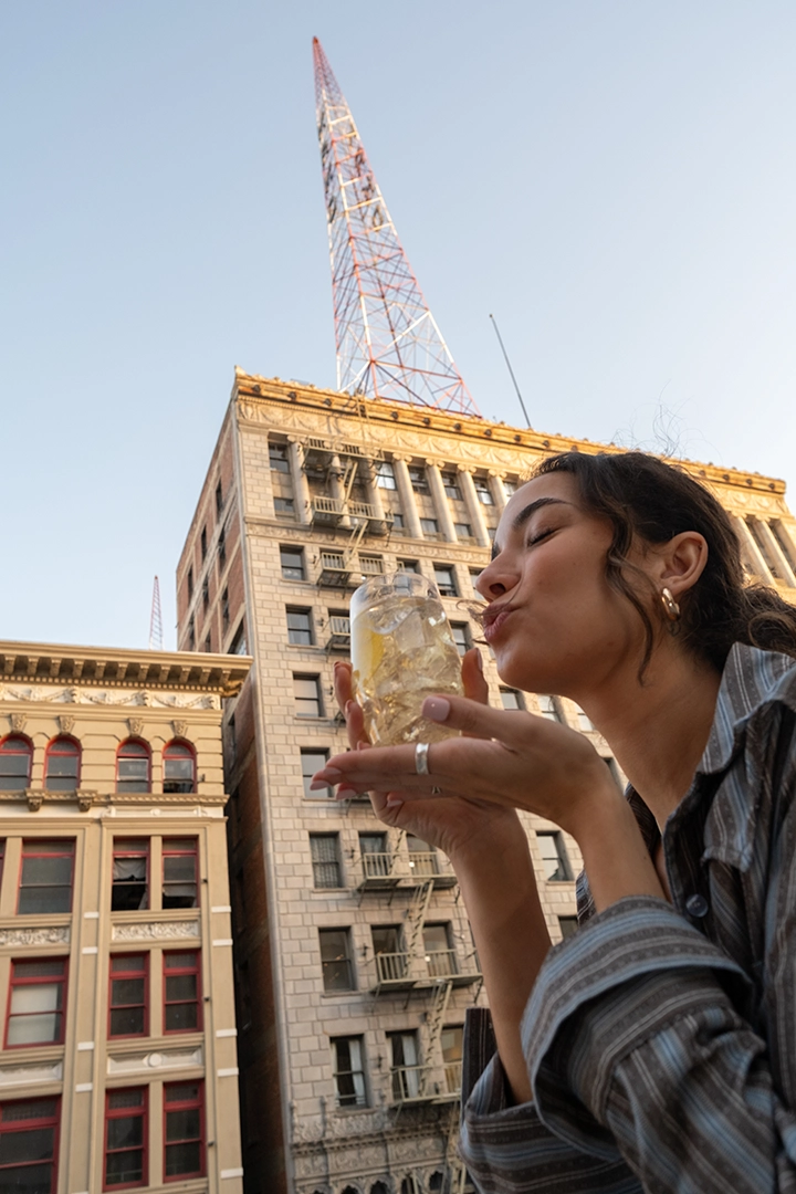 Nikaido Social 4 ‣ Merit/Andrew Woman in a striped shirt drinks iced lemon water from a glass on a city rooftop, with a tall radio tower and buildings in the background.